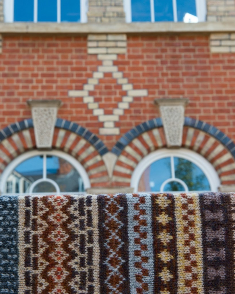Image shows stranded colour work swatch in the foreground of a picture of a wall featuring intricate brickwork
