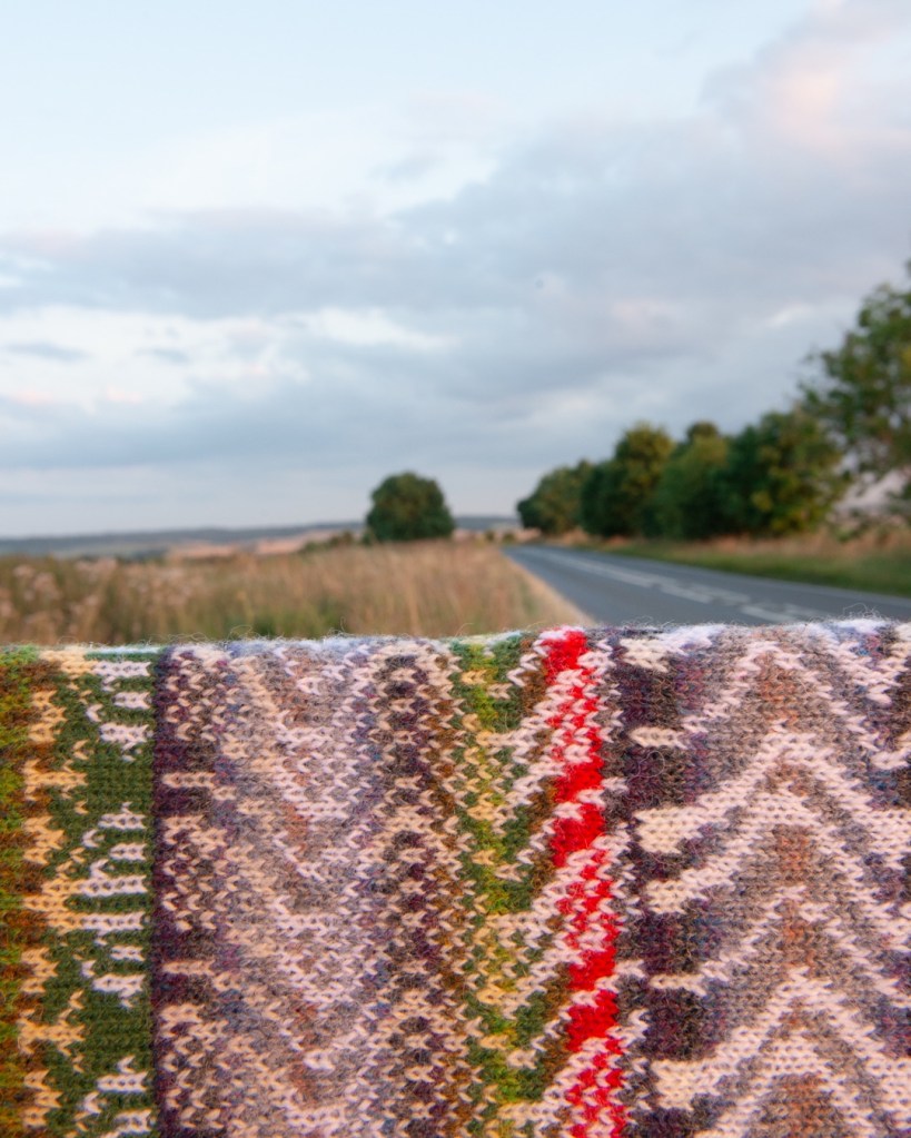 Image shows a stranded colourwork swatch in the foreground of a photo of a country road