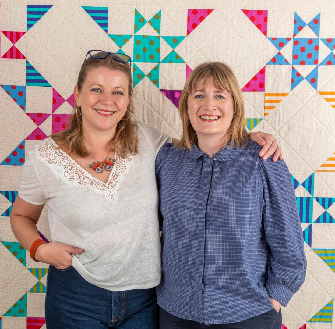 Image shows Jenni Smith & Kay Walsh standing together smiling at the camera in front of a beautiful patchwork quilt.