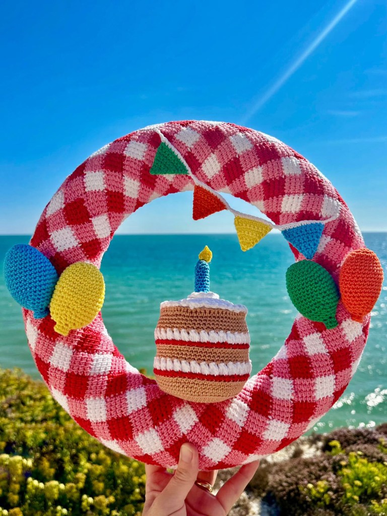 Image shows a Let's Celebrate Wreath held up in front of a view of the sea on a sunny day