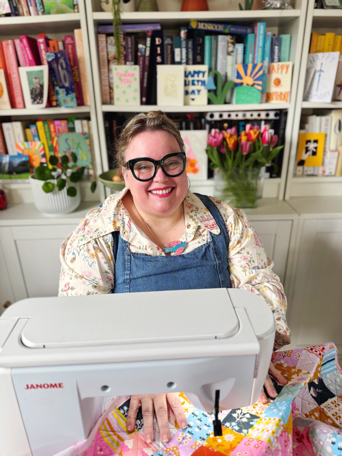 Image shows Kat Molesworth sitting at a table behind a white sewing machine. With a bookshelf in the background
